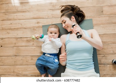 Top View Of Mother With Baby Boy Lying On Floor And Playing With Dumbbells 