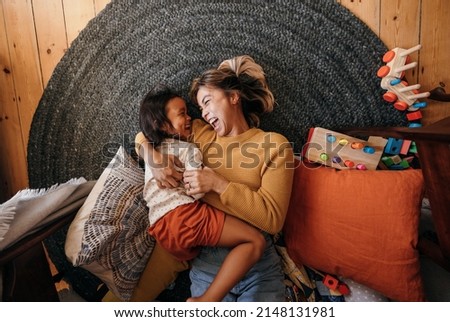 Top view of a mother and her daughter laughing cheerfully at home. Happy mother and daughter lying on the floor in their play area. Mother and daughter spending some quality time together.