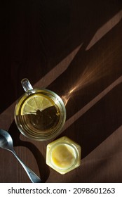 Top View Of A Mug Of Green Tea With Lemon, A Spoon And A Jar Of Honey On A Wooden Table With Shadows From The Sun.