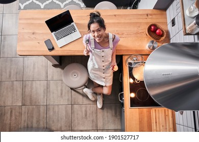 Top View Of Multiracial Woman Standing At The Kitchen With A Cooking Ladle Spoon While Eating Apple At Home. Woman Looking At The Camera And Smiling. Cooking Concept