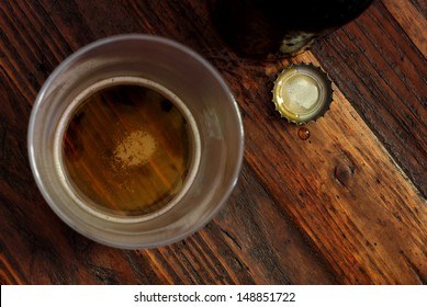 Top view of near empty beer glass with bottle cap and bottle on rustic wood background.  Low key still life with directional natural lighting for effect.  Selective focus on bottle cap and droplet.