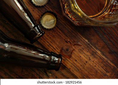 Top view of near empty beer glass with bottle caps and bottles on rustic wood background. Low key still life with directional natural lighting for effect. Selective focus on bottle caps and droplets. 