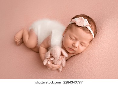 Top view of a newborn baby girl sleeping in a white jumpsuit with pink rabbit a white bandage and a pink flower on her head on a pink background. Beautiful portrait of a little girl 7 days, one week.