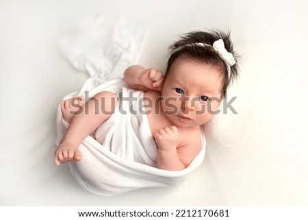Top view of a newborn baby girl lying in a white cocoon on a white bed with a white bandage. Beautiful portrait of a little girl with wide open eyes 7 days, one week. Photography in macro studio.