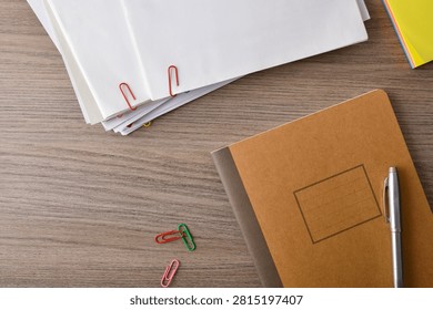 Top view of a notebook, stacked papers, paper clips and pen on a wooden desk with copy space, illustrating organization, office work and productivity.