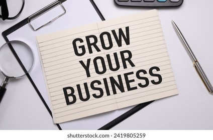 Top view of notepad with GROW YOUR BUSINESS text on wooden table, surrounded by calculator, magnifier, glasses, and pen, representing business concept and workspace.