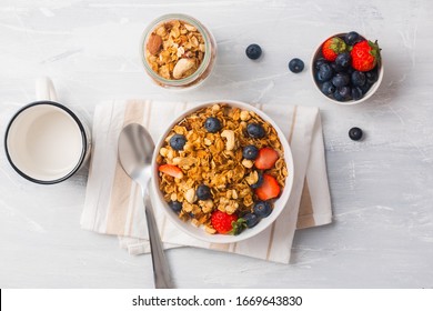 Top View Of Oats In A Cereal Bowl With Blue Berries And Strawberries In White Background On Top Of A Napkin. A Metal Spoon Of Oats, Small Bowl Of Oats And A Bowl Of Berries. A Coffee Cup At The Side.