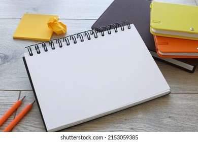 Top View Of Office Desk With Open Spiral Notebook, Orange Pencil And Green And Orange Notepad On Wooden Table