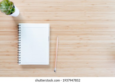 Top view of office desk table with open spiral notebook, pencil, and small tree in a white pot on wood table. Flat lay.