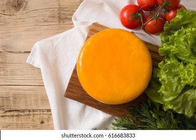 Top view on big cheese head in yellow vacuum package on wooden board served with tomatoes and fresh salad. Serving French homemade cheese. Food concept