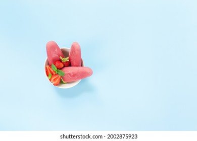 Top View On Bowl With Fruity Pink Frozen Sorbet Ice-cream On Plastic Stick With Strawberry, Mint On Blue Background. Frozen Mixed Fruits With Yogurt In A Form Of Ice-cream. Kids Love To Eat It A Lot