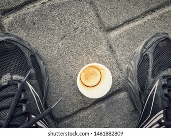 Top view on an empty paper cap of coffee standing on pavement between human feet wearing old sneakers