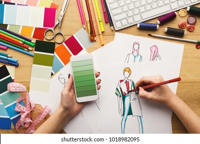 Top view on fashion designer at work. Female hands drawing clothes sketch at her creative workspace and using smartphone with green gradient color swatches on screen, top view