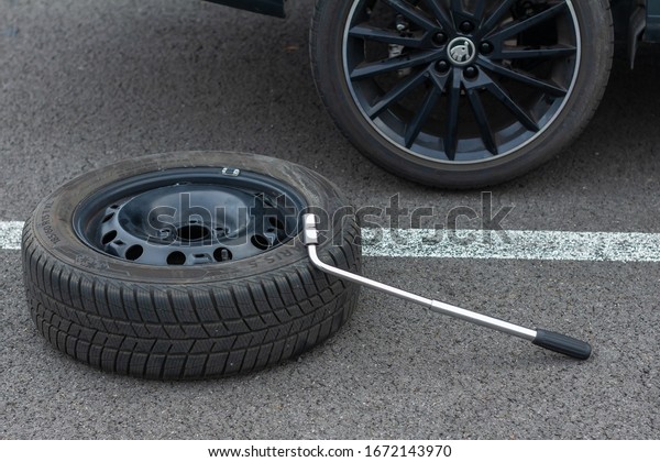 Top view on a flat tire car wheel and screwdriver\
are on a asphalt road on the broken car background. Jack is lifting\
up a vehicle. Automobile service. Tire replacement concept, Prague,\
March, 2020.