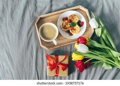 Top view on holiday breakfast on bed. Mother's day, 8 march, valentine's day concept. Delicious branch - coffee, cottage cheese, pancakes, gift box and fresh tulips - Powered by Shutterstock