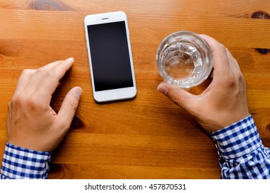 Top view on man working on mobile phone, hands and glass of water on wooden desk background.