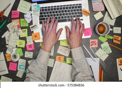 Top view on man working on laptop computer with notes all around his office desk. Overwhelmed with work concept.