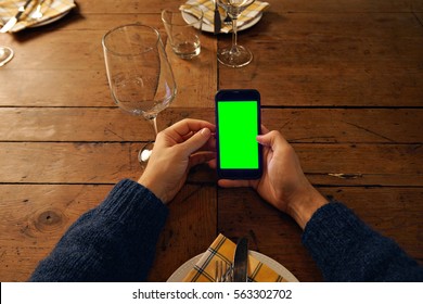 Top View On Man's Hand Using Smart Phone Above Served Table In Restaurant, Next To Empty Wine Glass. Screen Filled With Chromakey Green