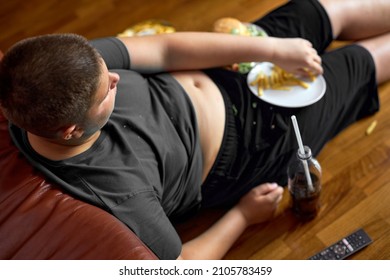 Top View On Obese Fat Overweght Boy Sitting On Floor Eating Fries, Overeating Teenager With Big Tummy Is Lazy, Watching Tv And Drinking Sweet Beverages At Home Alone, Need To Keep Diet