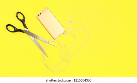 Top View On Rose Gold Cell Phone With Ear Buds Cable To Be Cut With Large Scissors On Yellow Background.