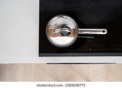 top view on stainless steel saucepan with lid on glass ceramic induction stove with temperature indicator and sensor buttons at kitchen, using modern household appliance