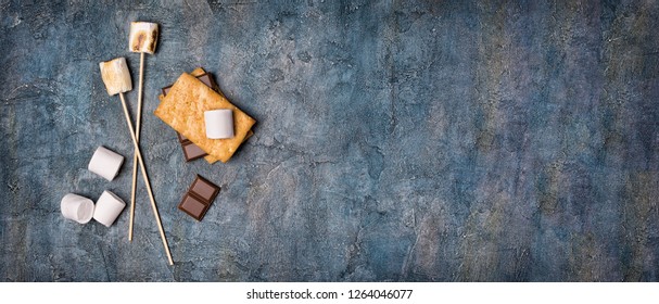Top view on sticks with roasted on campfire marshmallow, crackers and chocolate as ingredients for s'mores on blue concrete background with copy space