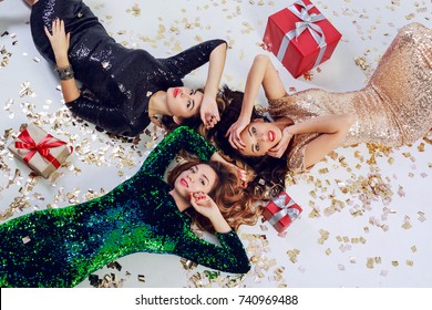  Top View On Three Gorgeous Girls Lying On The Floor, Celebrating New Year Or Birthday Party. Wearing Luxury Sequins Dress  And Jewelry. Golden Shining Confetti , Red Gift Boxes. 