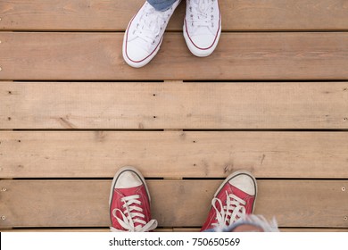 Top view on two people's feet in white and red sneakers standing on wooden floor or bridge.