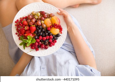 Top View On Woman Sitting Cozy On The Couch And Enjoying Delicious Summer Fruit And Berries. Girl Holding White Plate With Apricots, Raspberries, Strawberries, Blackcurrant, Blueberries On Her Knees