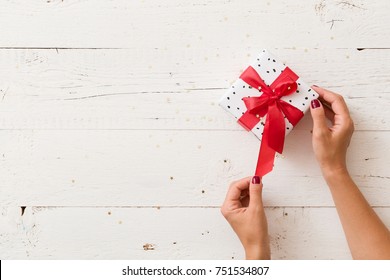 Top View On Woman's Hands Unwrapping Christmas Or Birthday Gift Box With Red Satin Ribbon On White Wooden Background.