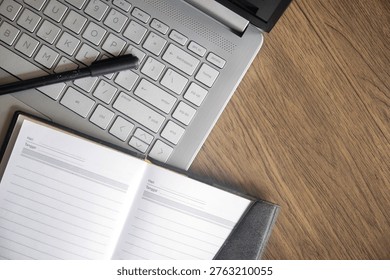 Top view of open blank notebook with black pen resting on silver laptop keyboard over dark wood grain desk. Concept for planning and remote work.