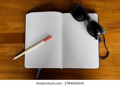 Top view of an open blank notebook with pen and sunglasses on a wooden table. Minimal lifestyle workspace scene with copy space, summer mood, journaling, travel planning, and productivity concept.