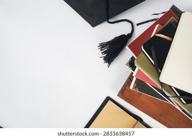 Top view of an open blank notebook with brown pages beside a stack of books, black graduation cap and tassel on a white background.