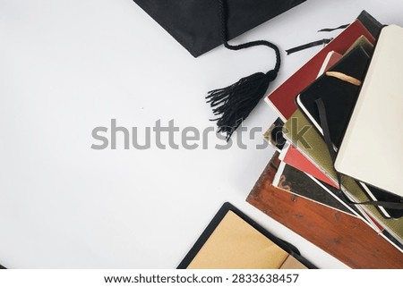 Top view of an open blank notebook with brown pages beside a stack of books, black graduation cap and tassel on a white background.