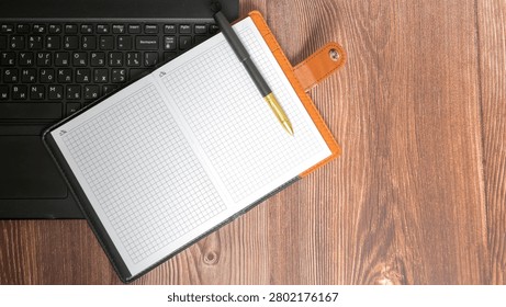 top view of an open checkered notebook with a pen lying on the keyboard of a black laptop on a wooden brown table on the right side of the frame close up