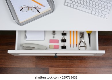 Top View Of An Open Desk Drawer Showing The Organized Items Inside. The Top Of The Desk Has A Computer Keyboard And Wire In-box With Paper And Pencil. The Drawer Has Pencils, Erasers, Stapler And More