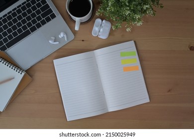 Top view open notebook, wireless earphones, laptop and coffee cup on wooden table.