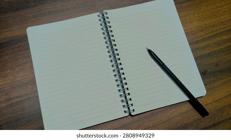 Top view of an open wire-bound notebook with lined pages on a dark wooden table. A black pen rests on the right-hand page. The left page is blank. Empty, clean, ready to write.