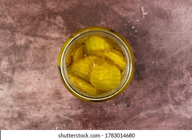 Top View Of An Opened Glass Jar Filled With Hamburger Dill Pickle Slices On A Maroon Tabletop Illuminated With Natural Lighting.