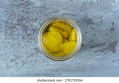 Top View Of An Opened Glass Jar Filled With Hamburger Dill Pickle Slices On A Gray Tabletop Illuminated With Natural Lighting.