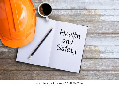 Top View With Orange Safety Engineer Helmet And Text Health And Safety On Notebook And Cup Of Coffee On Wood Table Background Or Office Desk Background.