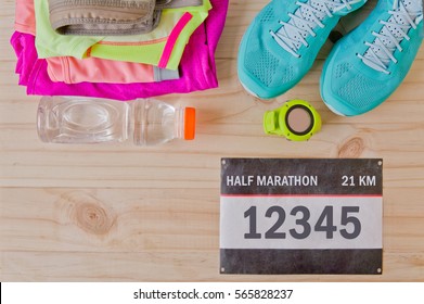 Top View Of Outfit For Mini Marathon Runner On Wooden Background: Bib Number, Gps Watch, Running Shoes, Shorts, Shirt, And Sport Bra. 