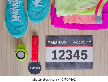 Top View Of Outfit For Runner On Wooden Background: Bib Number, Finisher Medal, Gps Watch, Running Shoes, Shorts, Shirt, And Sport Bra. Horizontal Orientation.