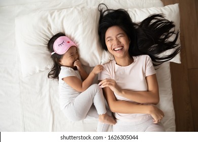 Top view of overjoyed young Asian mother lie in comfortable bed with small biracial girl child have fun in bedroom together, happy Vietnamese mom and little daughter laugh enjoy early morning at home