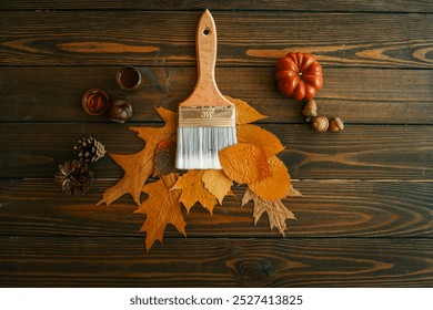 Top view of a paintbrush placed on dried autumn leaves with acorns, pine cones, and a pumpkin on a rustic wooden background. - Powered by Shutterstock