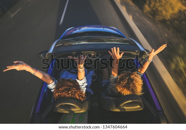 Top view a pair of euphoric women in a\
convertible car twisting and waving. Two curly girls on vacation\
having fun driving the auto, laughing happily singing, enjoying\
freedom in a beautiful\
sunlight