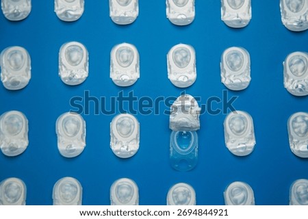 Top view of the pattern in focus is an open container with contact lenses on top of which lies a transparent blue lens against the background of containers with contact lenses on a blue background