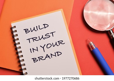 Top View Of Pen, Magnifying Glass And Notebook Written With BUILD TRUST IN YOUR BRAND. Business Concept.