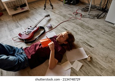 Top view of pensive male musician and songwriter thinking about new song. Domestic hobby, entertainment and leisure. Young creative european man lying on floor with papers and electric guitar at home