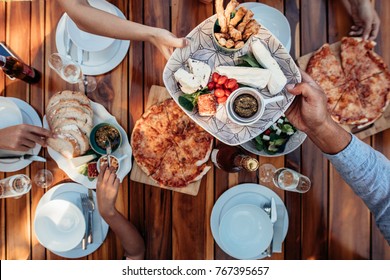 Top View Of People Eating Meal At Table Served For Party. Friends Celebrating Housewarming Party On Wooden Table. Male Hands Passing Food Plate To Female Guest.